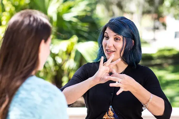 Female student with blue hair (facing camera) signs to second female student (not facing camera).