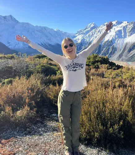 Katie Kress poses for an excited photo with her arms in the air. A mountain range can be seen behind her.