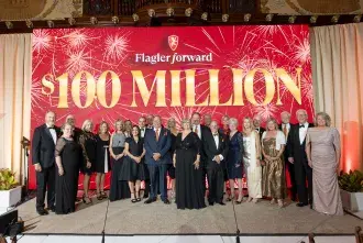 A group shot of Flagler College President Delaney and friends in front of a large banner that reads 'Flagler Forward $100 Million' at the celebration gala.