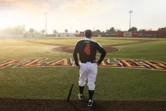 Coach Barnett facing away from the camera, leaning on a baseball bat an looking at an empty baseball field.
