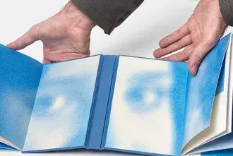 Visual artist and writer, Pablo Vindel, holds an open book where the pages are covered with speckles of blue paint that resemble a face.