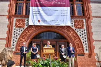President Delaney behind a podium on the steps of Ponce Hall under a large sign displaying the number twelve million