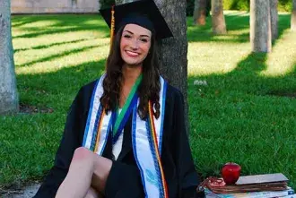 Sasha Pabis, wearing her graduation gown, sits on the grass in the Flagler Palm Garden