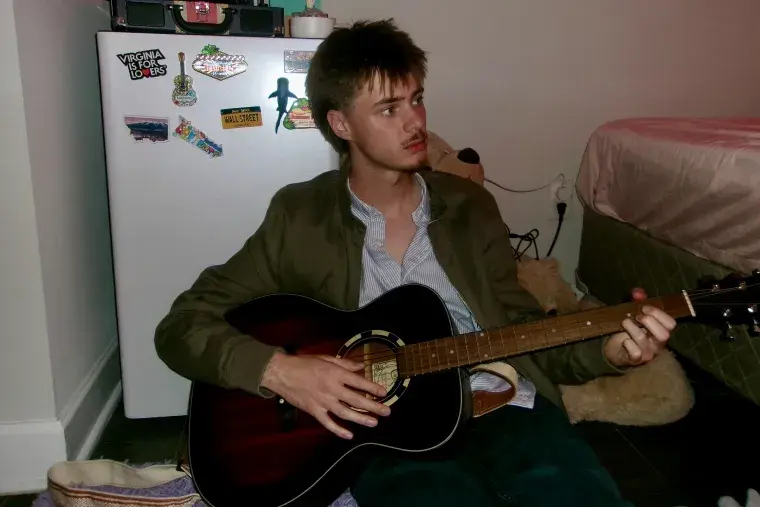 Student Musician Ben Baine plays his guitar while sitting on the floor in his bedroom