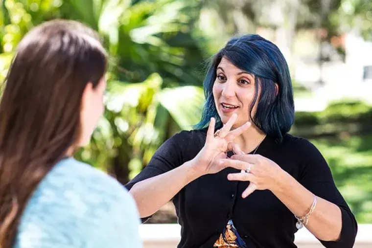 ASL_Lab.jpg Female student with blue hair (facing camera) signs to second female student (not facing camera).