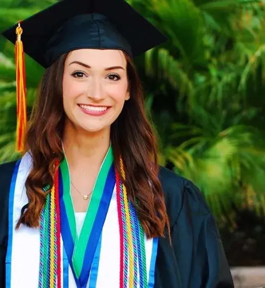 Sasha Pabis, wearing her graduation gown, stands at the gated entrance to Flagler College