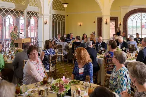 Alumni sit around circular tables in the Ponce Hall solarium. Woman speaks from behind a podium. 