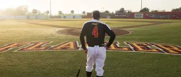 Coach Barnett facing away from the camera, leaning on a baseball bat an looking at an empty baseball field.