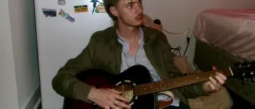 Student Musician Ben Baine plays his guitar while sitting on the floor in his bedroom