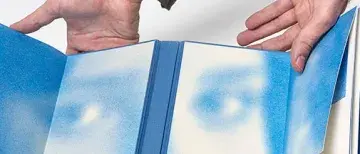 Visual artist and writer, Pablo Vindel, holds an open book where the pages are covered with speckles of blue paint that resemble a face.