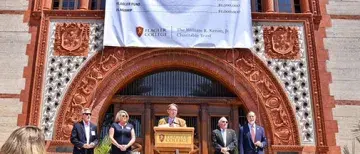 President Delaney behind a podium on the steps of Ponce Hall under a large sign displaying the number twelve million