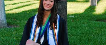 Sasha Pabis, wearing her graduation gown, sits on the grass in the Flagler Palm Garden