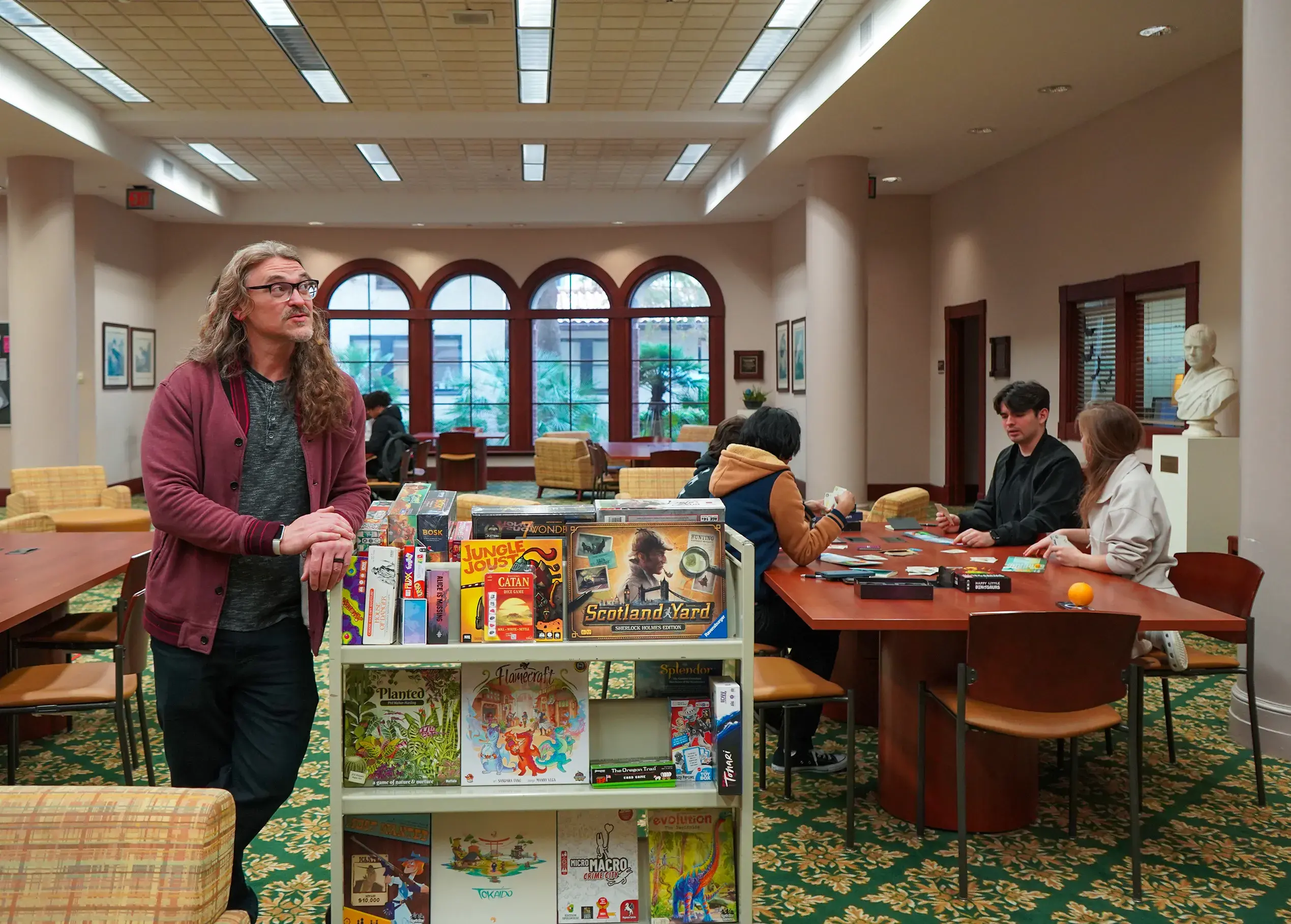 Professor Moody posing with his "game cart" as students sit at table playing game behind him